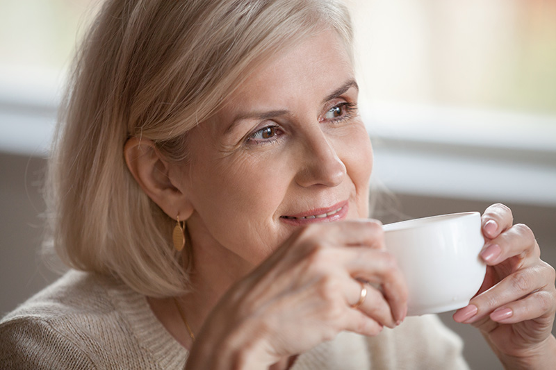 Glückliche Seniorin mittleren Alters, die entspannt eine Teetasse hält und lächelnd zur Seite blickt.